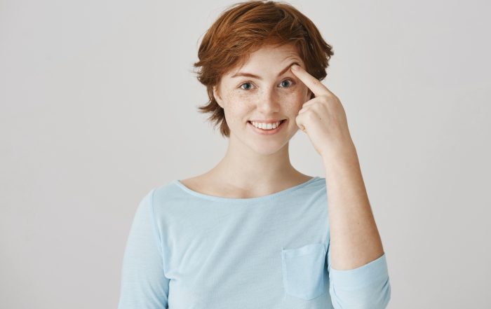 Girl can not lift her eyebrow without help of hand. Portrait of happy attractive redhead female student with freckles in ordinary clothes pulling brow with index finger, smiling and being in good mood