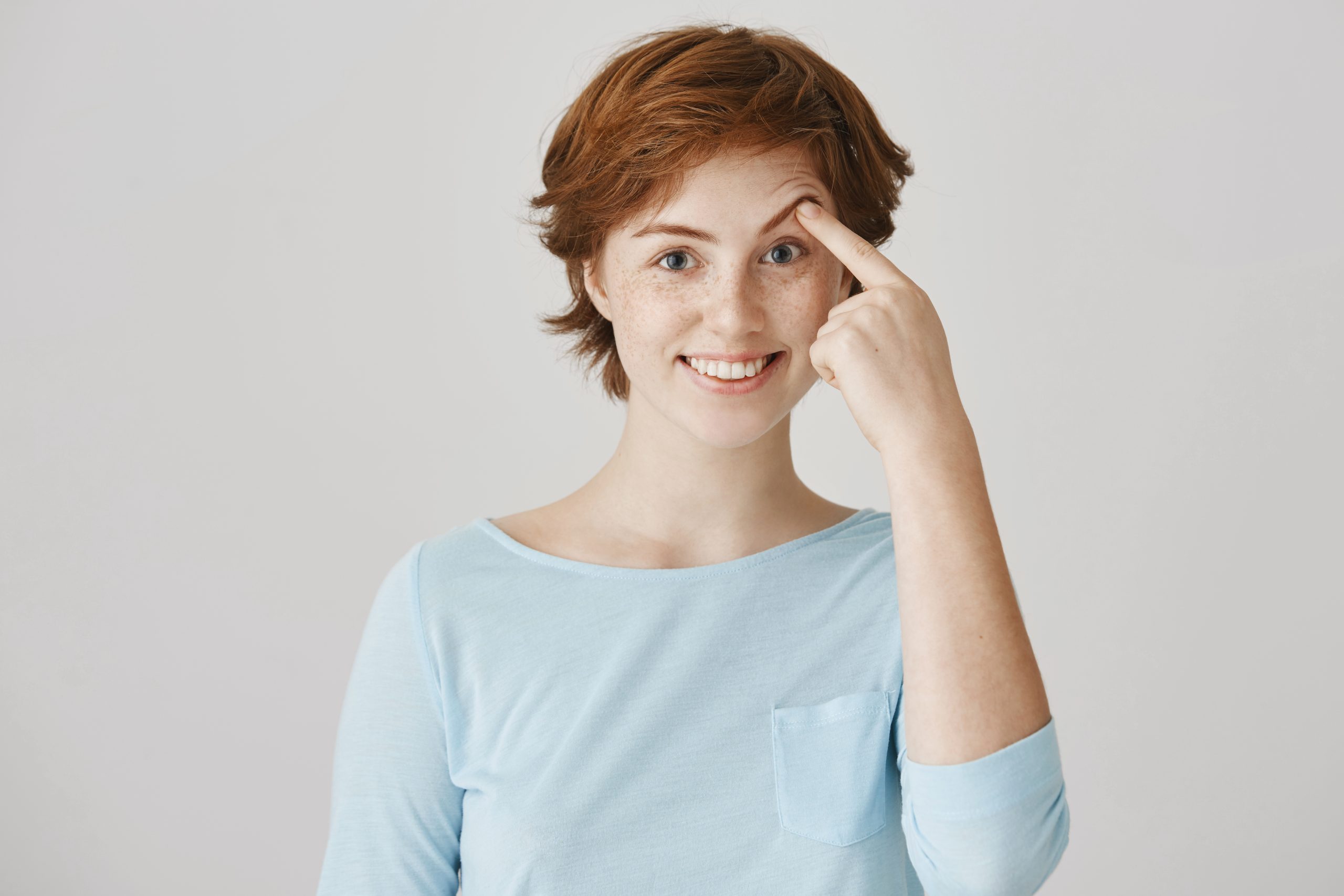 Girl can not lift her eyebrow without help of hand. Portrait of happy attractive redhead female student with freckles in ordinary clothes pulling brow with index finger, smiling and being in good mood