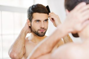 Morning routine. Rear view of handsome young beard man combing his hair while standing against a mirror