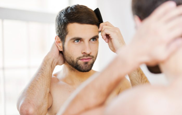 Morning routine. Rear view of handsome young beard man combing his hair while standing against a mirror