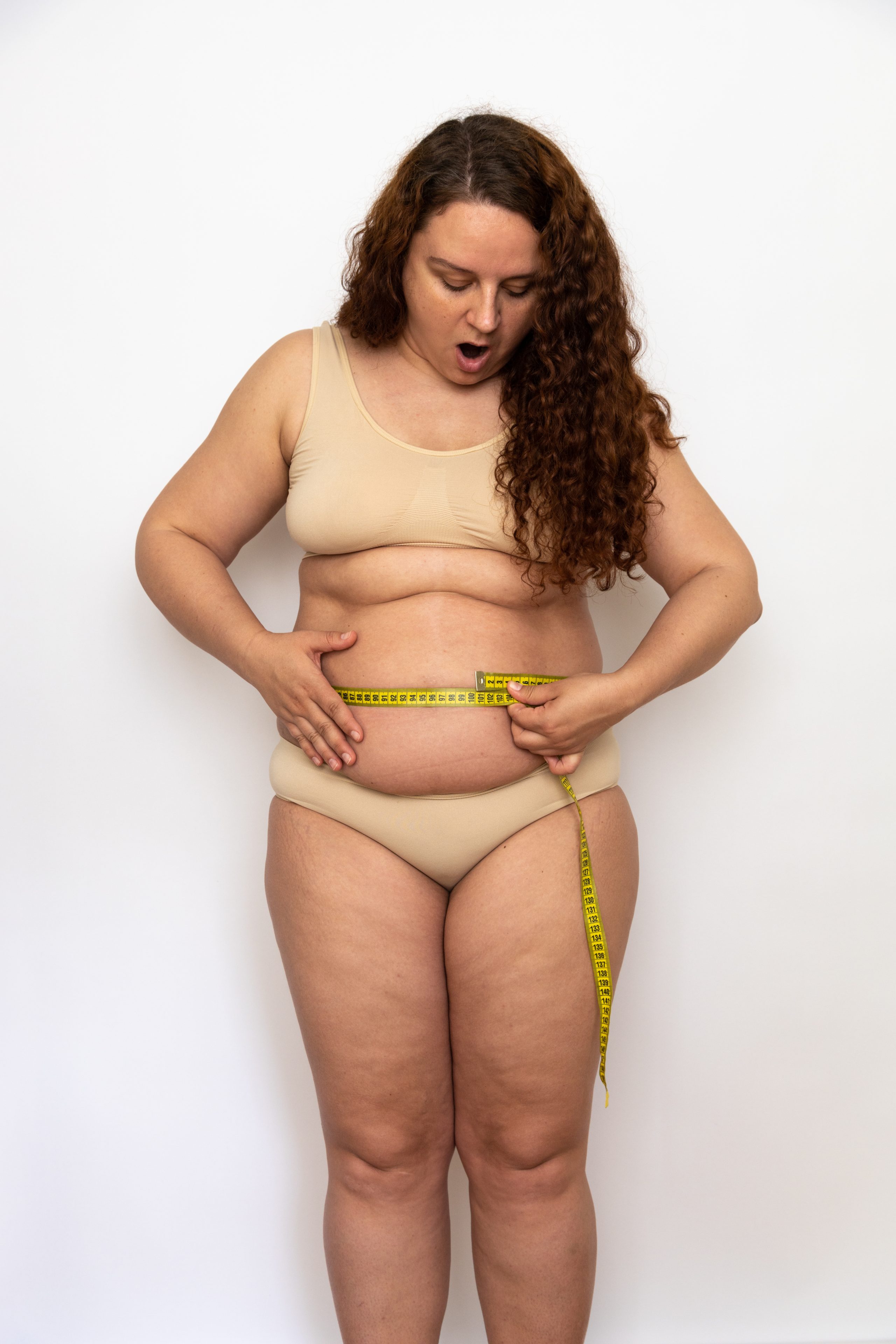 Portrait of shocked plump middle aged woman with curly hair wearing beige underwear, measuring waist with yellow tape.