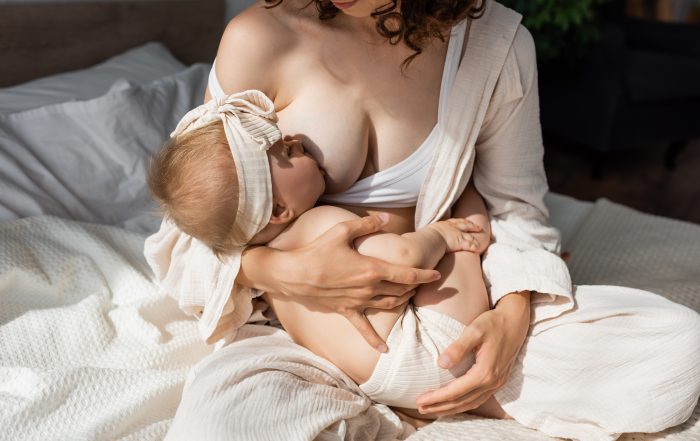 curly woman in loungewear sitting on bed and breastfeeding infant daughter