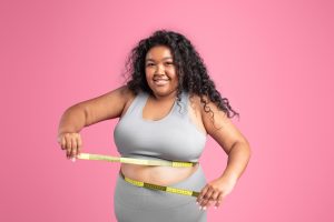 Smiling black overweight woman in sportswear checking waist with measuring tape, isolated on pink studio background