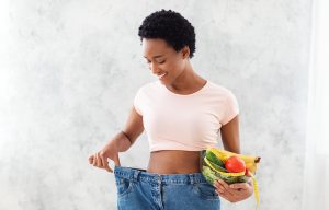Happy African American woman in old oversized jeans holding bowl with fruits and vegetables on grey background