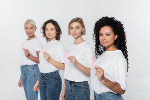Interracial women holding pink ribbons of breast cancer awareness and looking at camera isolated on
